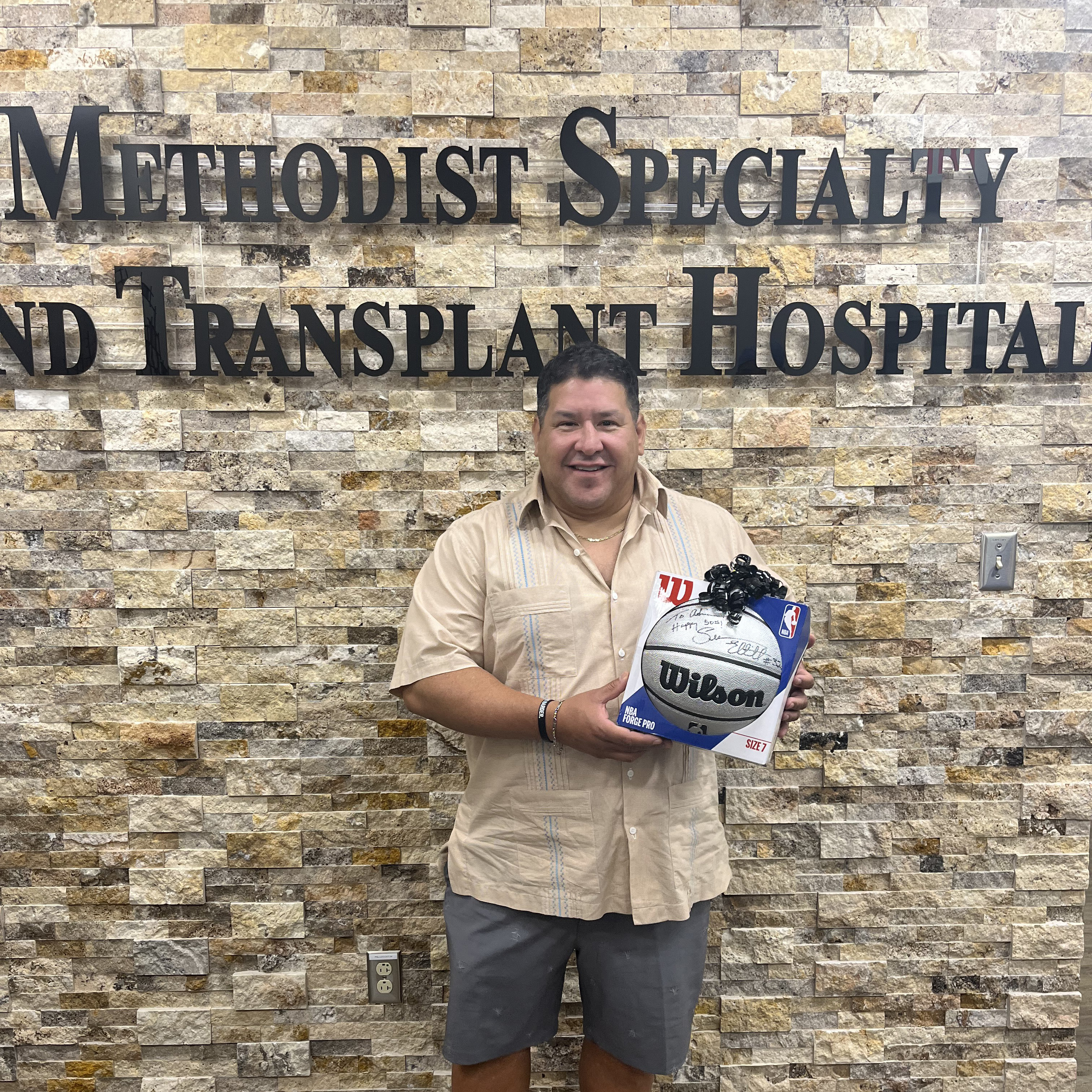 Adrian Lozano standing in front of the Methodist Hospital Specialty and Transplant sign holding a signed basketball 