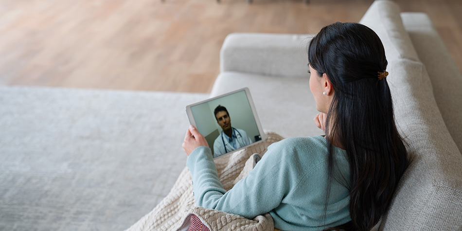 A woman sitting on a couch with a blanket. She's holding a tablet showing a medical professional on the screen in a video chat.