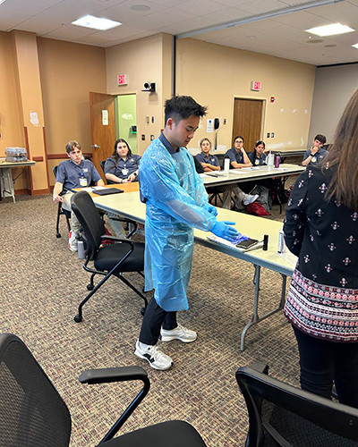 A volunteer adjusts their blue medical gown and gloves in a demonstration in front of other seated volunteers.