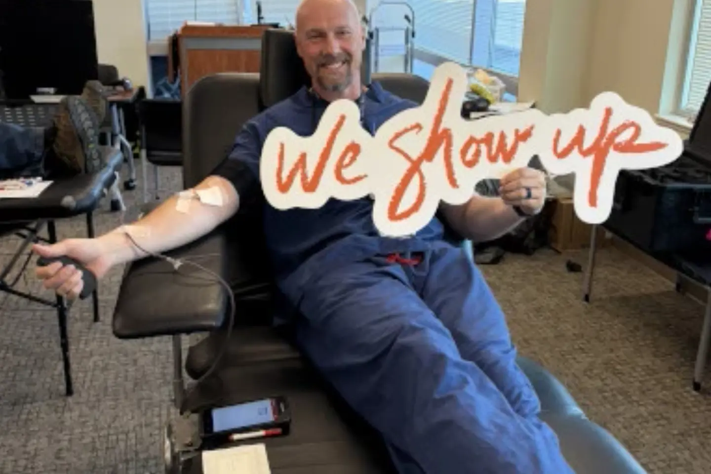 A West Valley Medical Center employee smiles while donating blood, holding a sign that says, "We Show Up®."