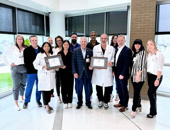 A group of medical professionals are standing in a hospital lobby holding a plaque. The group photo was taken to celebrate the signature status.