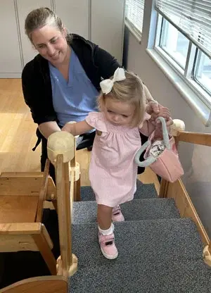 A medical professional smiles while helping Emma Mansour walk up stairs during physical therapy.