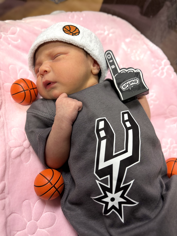 Newborn baby dressed in Spurs-themed outfit lying next to a mini basketball at a Methodist Healthcare hospital.