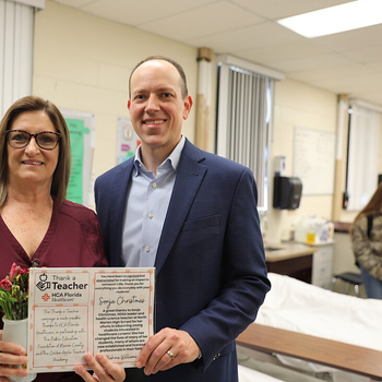 A woman in maroon scrubs and a man in a blue suit stand side by side in a healthcare classroom, smiling at the camera while holding a "Thank a Teacher – HCA Florida Healthcare" certificate and flowers. A hospital bed and clinical training supplies are visible behind them.