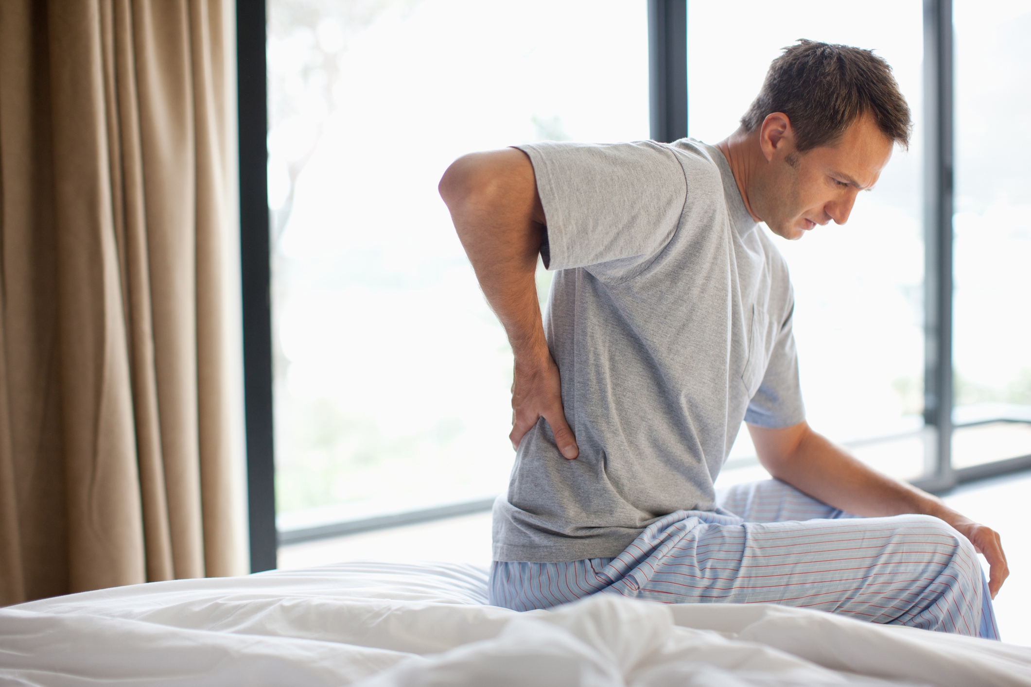 Man sits on his bed in pajamas, clutching his back in pain.