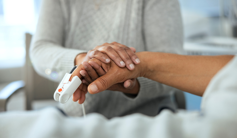 The closeup of holding hands. An adult patient with a thermometer on their finger and a child standing beside the hospital bed, holding the patient's hand.