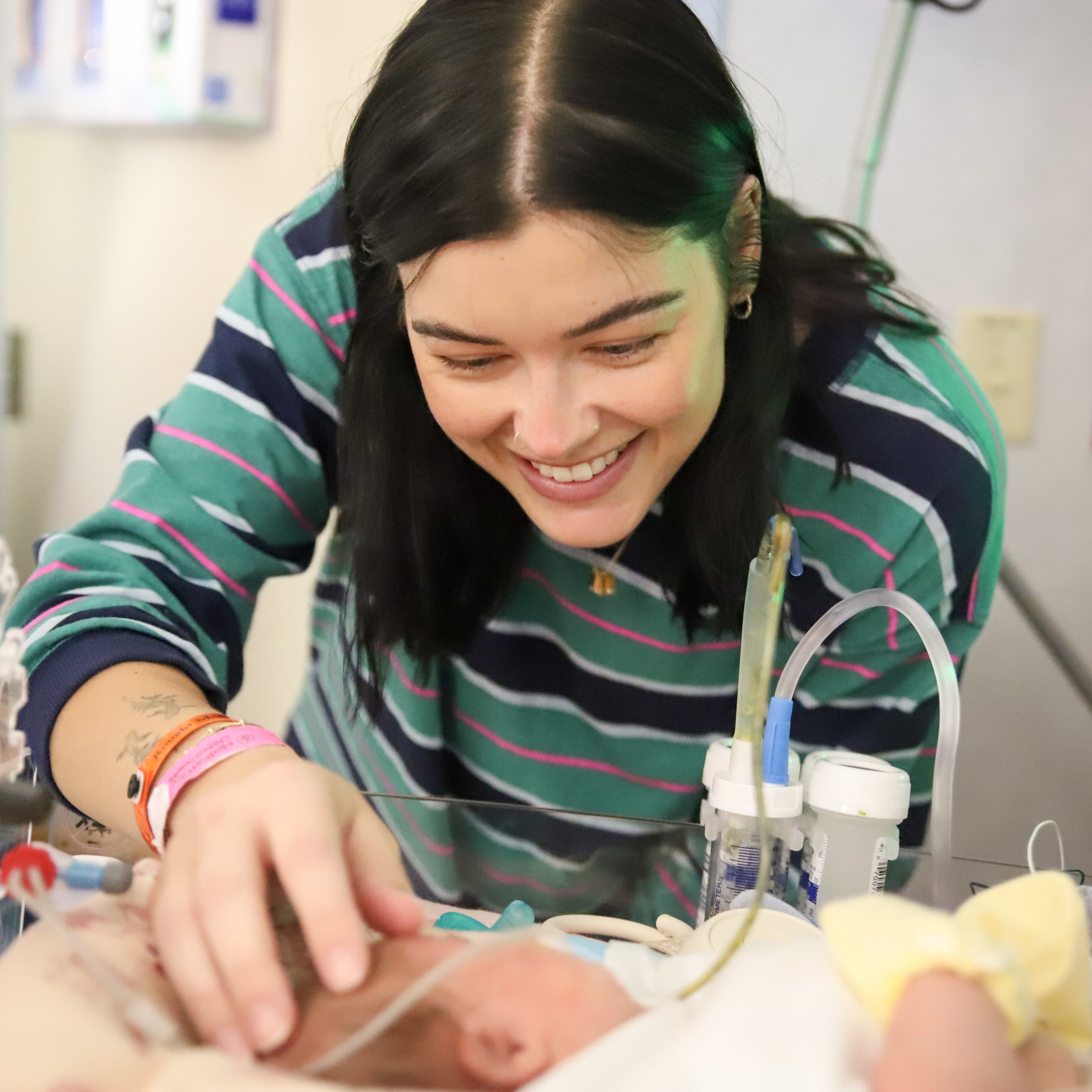 A smiling young woman with dark hair gently touches and looks at a newborn baby lying in a hospital incubator, surrounded by medical tubes and monitoring equipment.