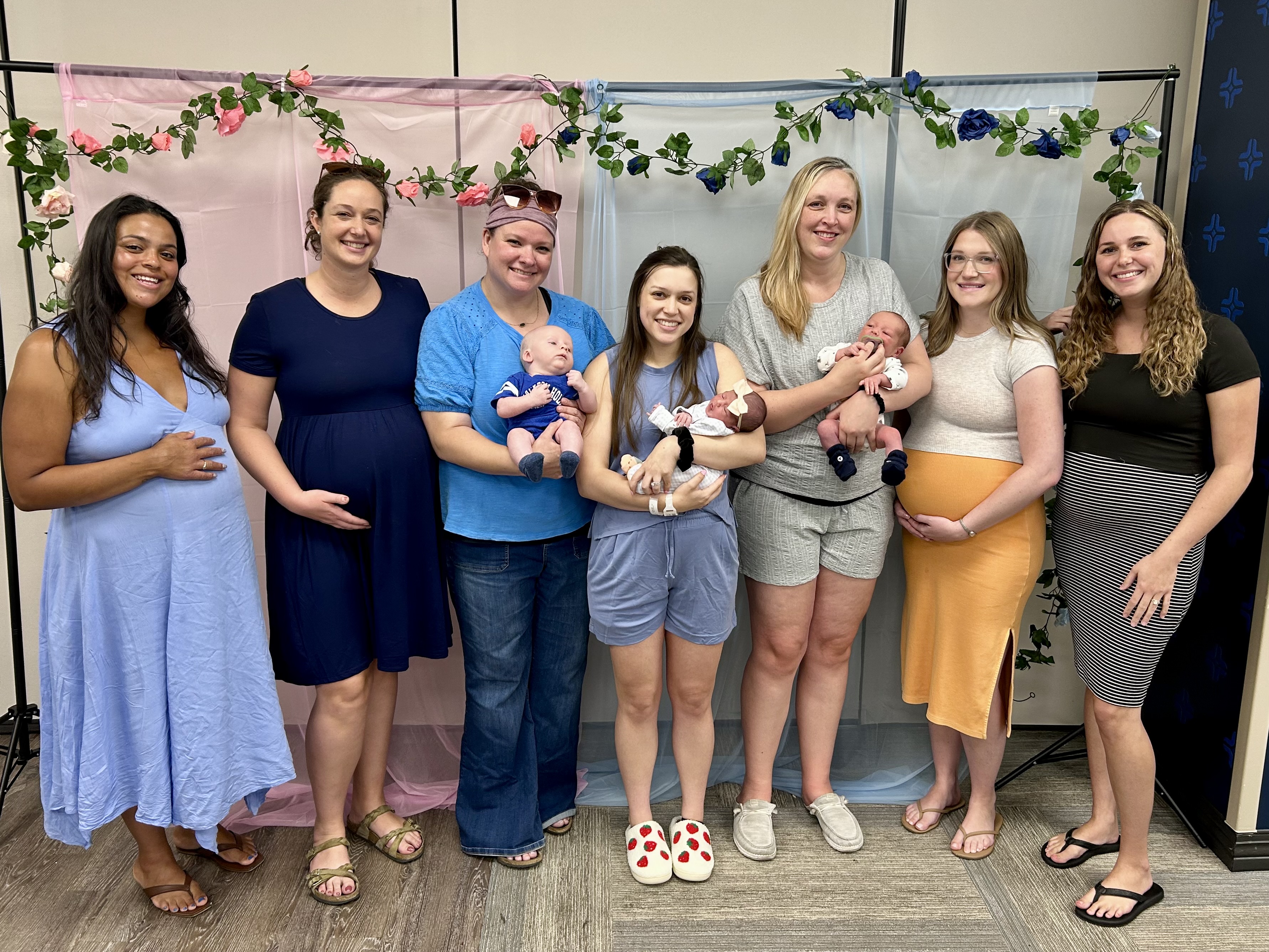 Group photo at a baby shower with 7 adults, including expectant parents and adults holding newborns, standing in front of pink and blue floral decorations.