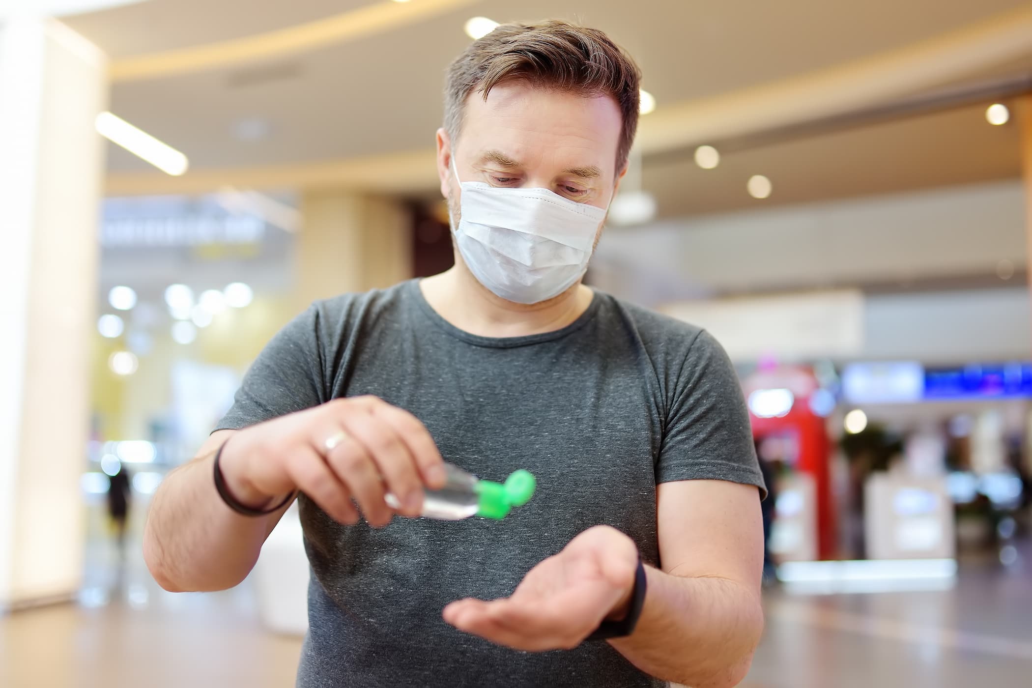 Male traveler wearing a mask applies hand sanitizer to his palm in an airport.