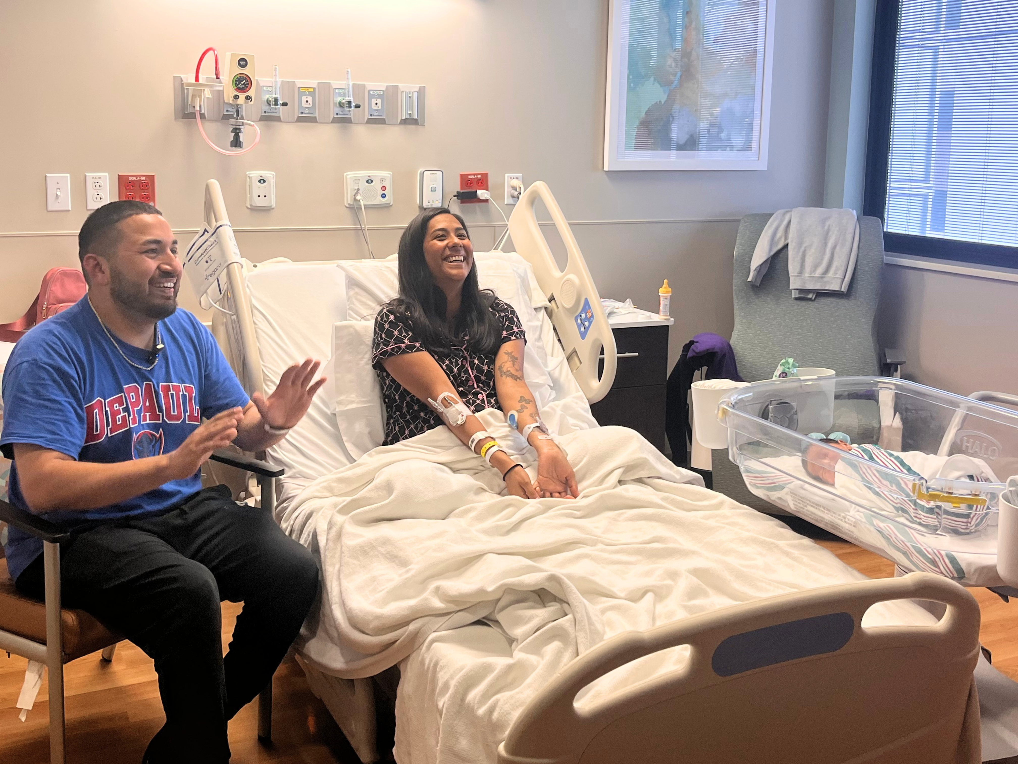 A smiling mother sits in a hospital bed beside the father of her newborn at Methodist Hospital Metropolitan. Their baby is resting in a bassinet next to the bed.
