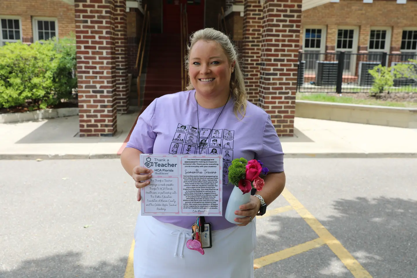 A woman in a purple T-shirt and white skirts holds a certificate and a small bouquet of flowers.