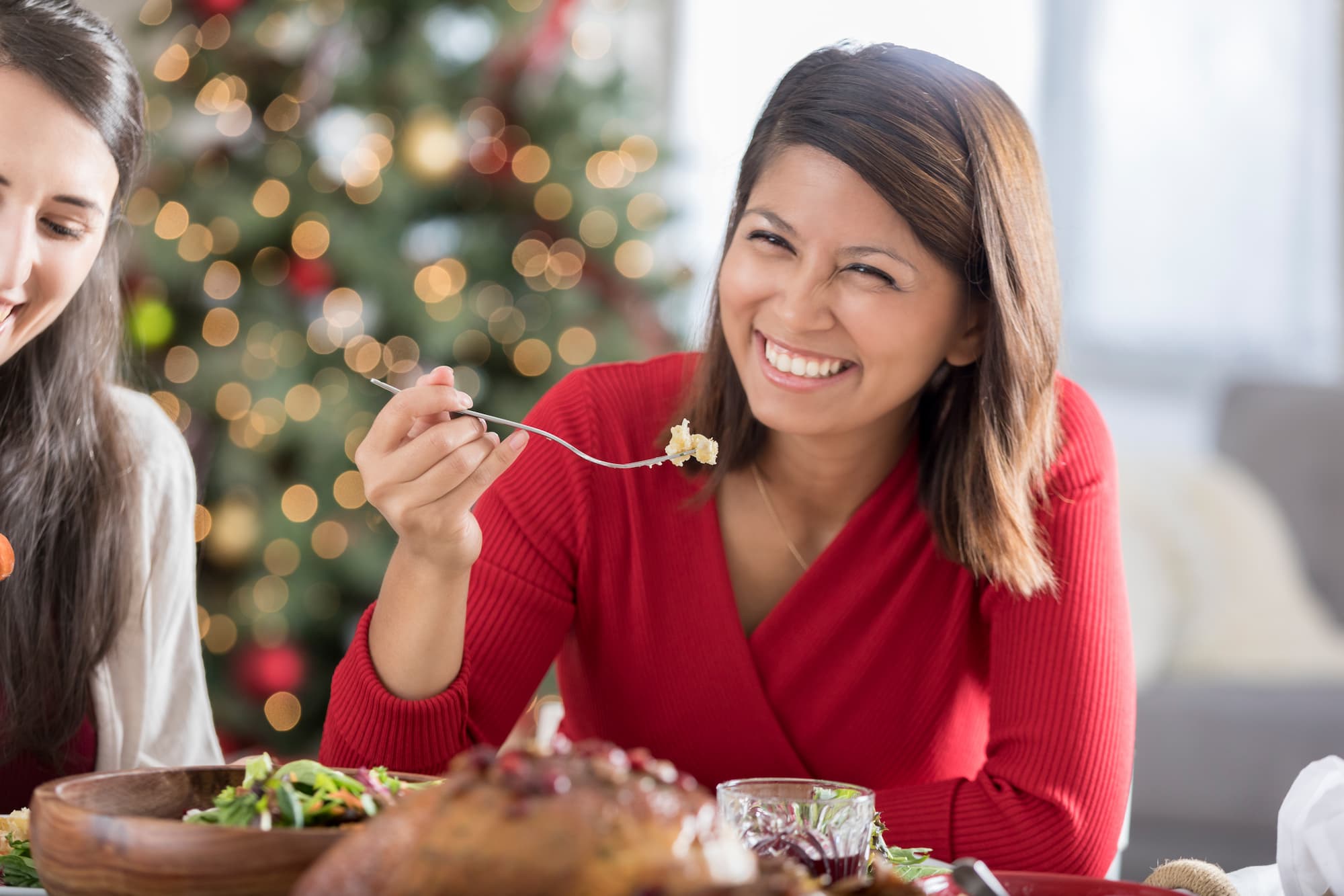 A woman smiles while holding her fork near her mouth as she is seated at a table during a holiday meal.