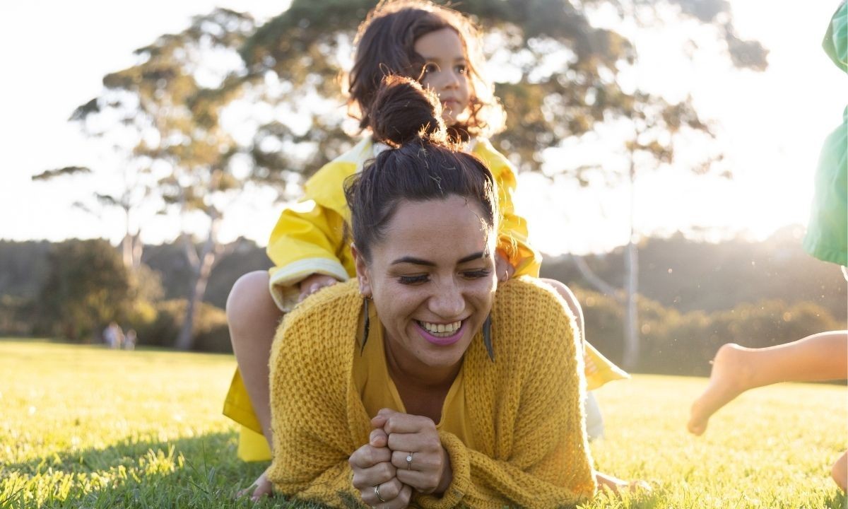 A woman lying on her belly on the grass, laughing joyfully as her young daughter sits on her back.
