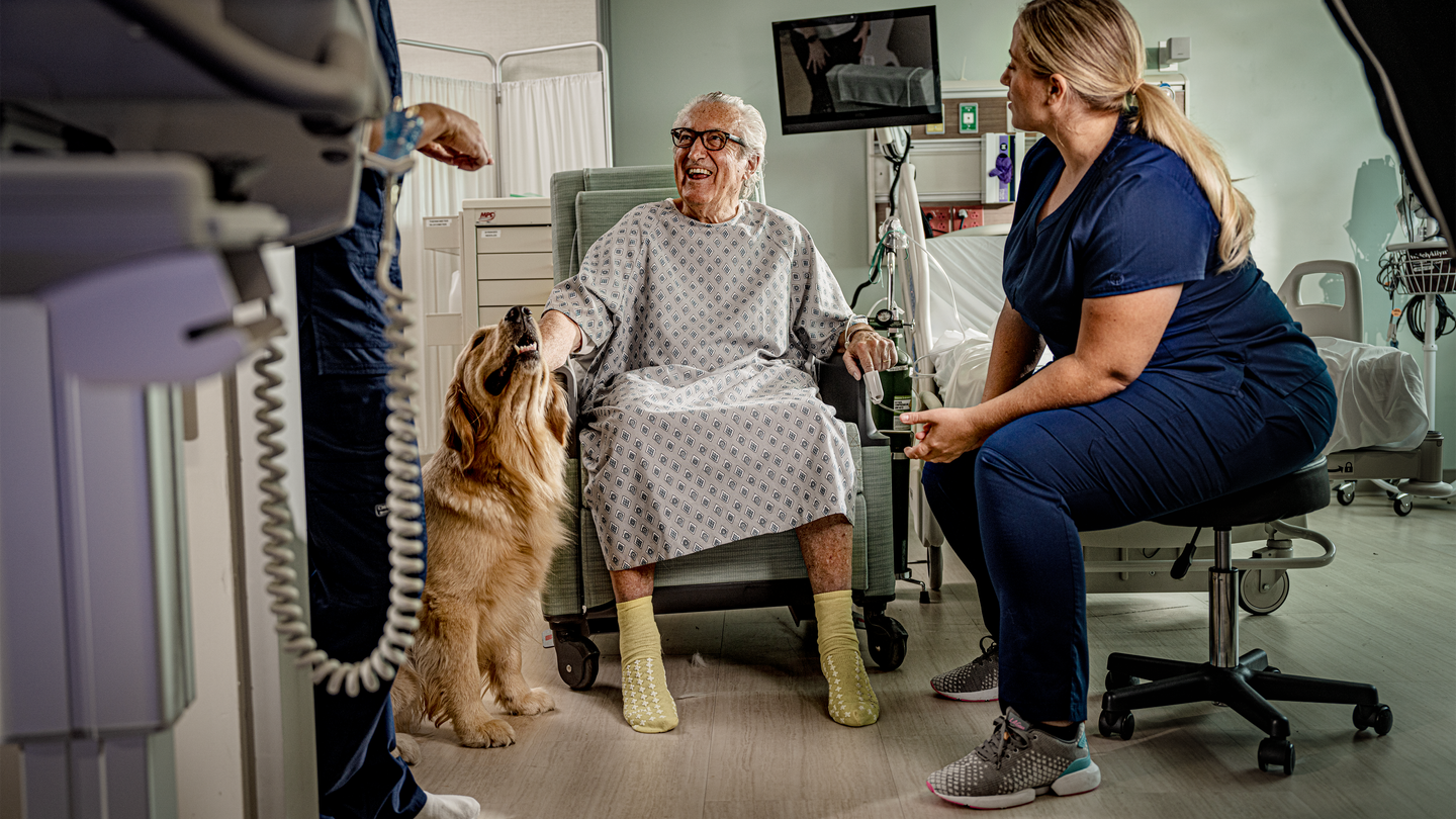 Nurses smiling and talking to a patient while he sits in a chair smiling while petting a therapy dog.