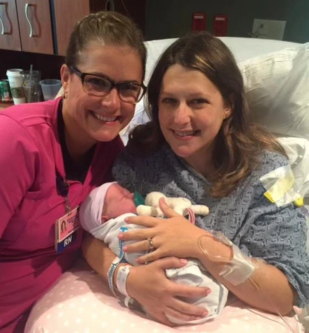 Female nurse wearing glasses and a pink scrub top, smiling next to a new mother wearing a hospital gown sitting in the bed holding newborn.