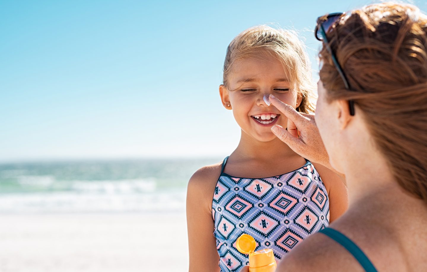 A girl smiles while a woman puts sunscreen on the tip of her nose at the beach.