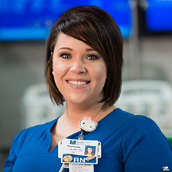 Stephanie Wassum smiles while wearing blue hospital scrubs and her hospital badge.