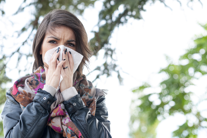 A brunette woman with black jacket and colorful scarf sneezes into a tissue, in the park.
