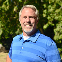 Bert smiles while standing outside, wearing a blue stripe polo shirt.