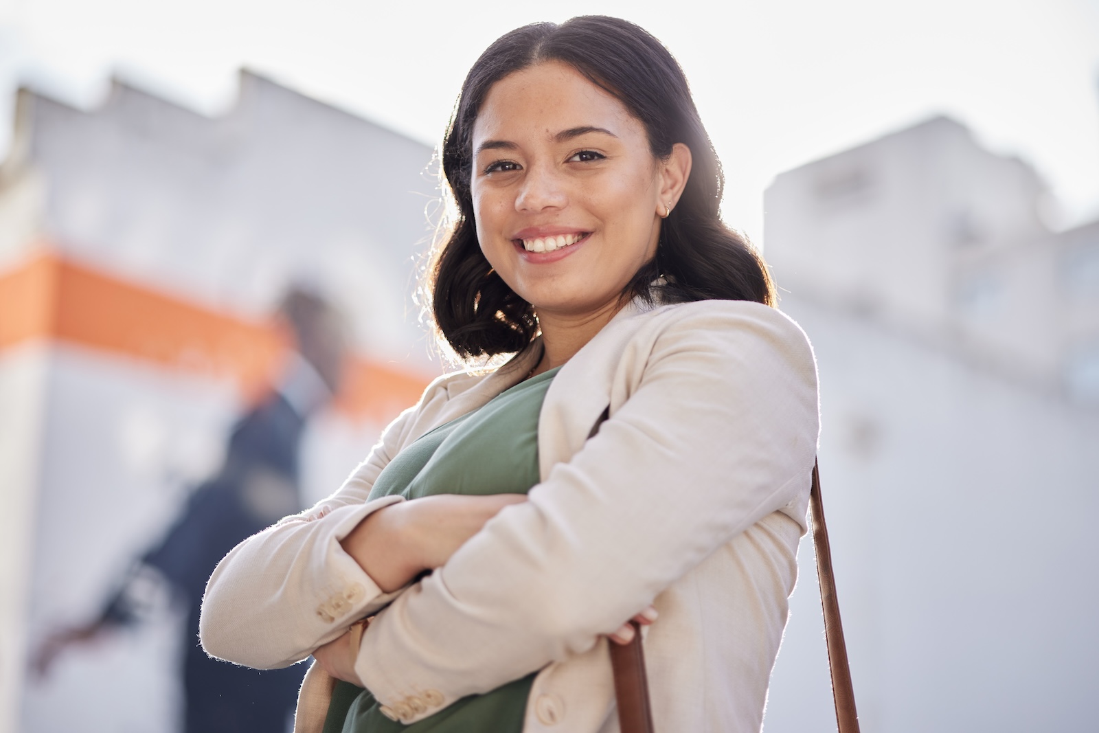 A woman smiles and crosses her arms while wearing a beige blazer and green shirt.