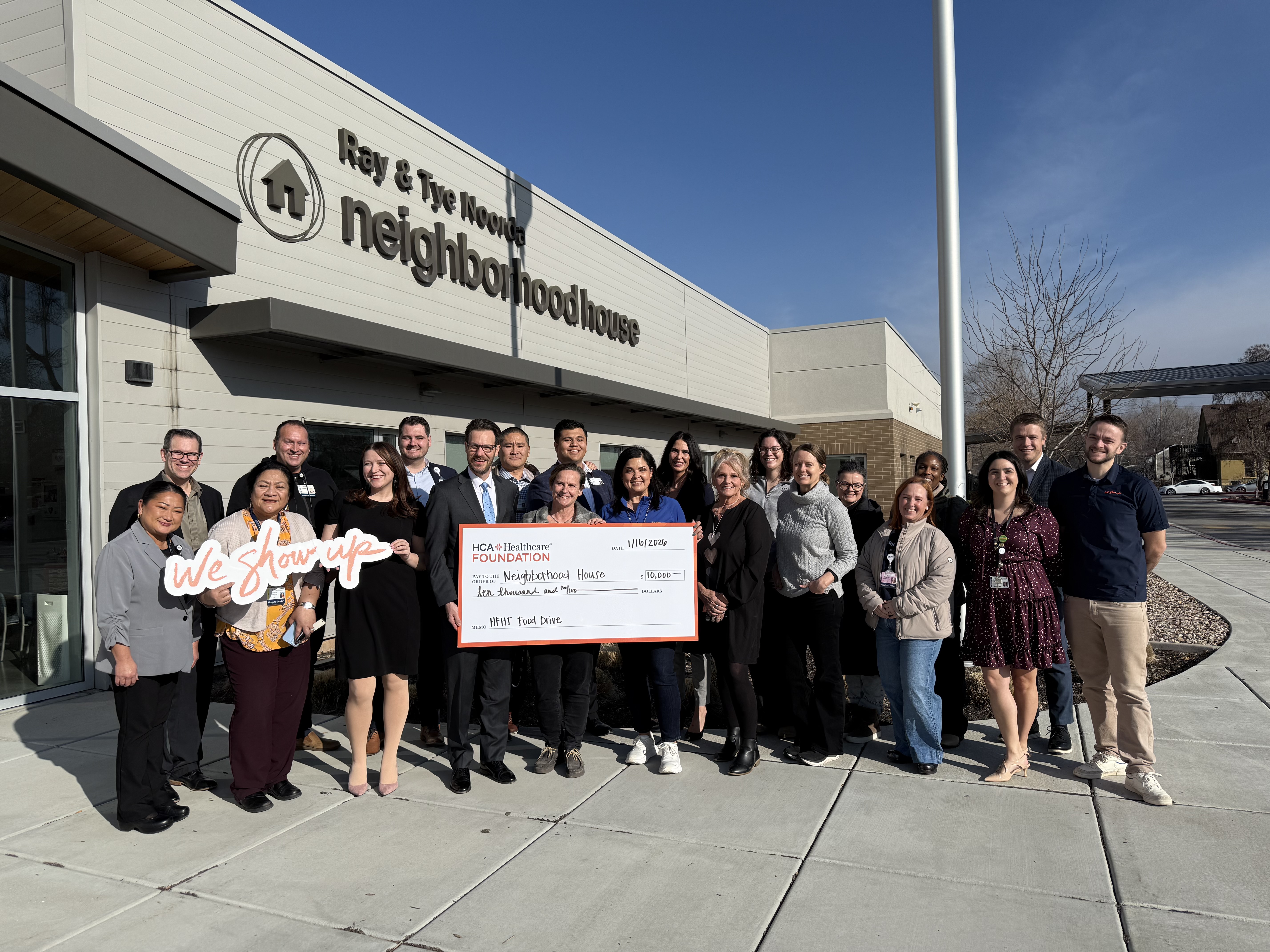 Members of the administration team at St. Mark's Hospital pose together in a group, smiling, alongside representatives of 501(c)3 local non-profit organization, Neighborhood House, outside its entrance. The group holds a large check made out to the non-profit worth $10,000 and a cut-out that reads, "We Show Up."