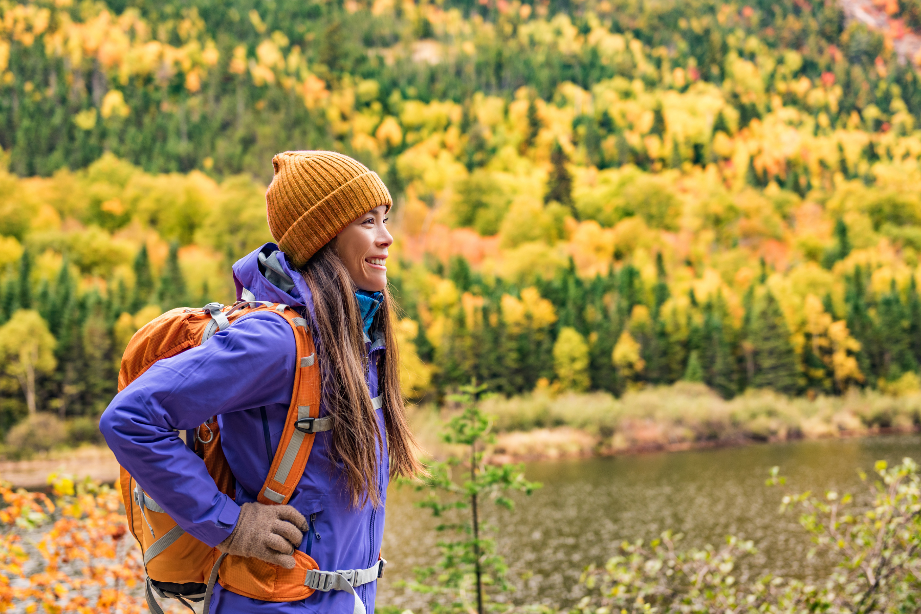 A woman wearing an orange beanie and backpack smiles while standing in front of a lake, with green and yellowing trees in the background.