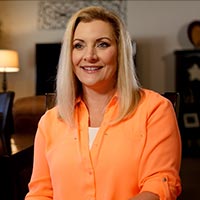 Brenda smiles while sitting in her living room, wearing an orange blouse.