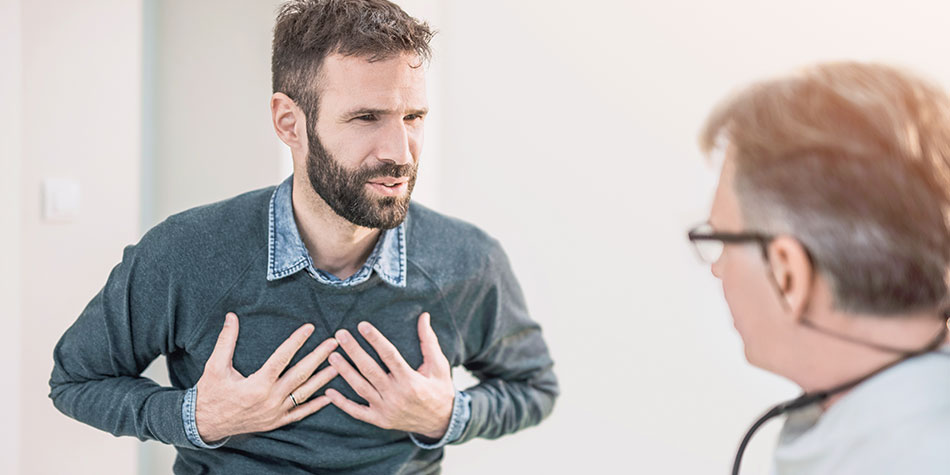 A bearded man holds his chest in discomfort and talks to male doctor 