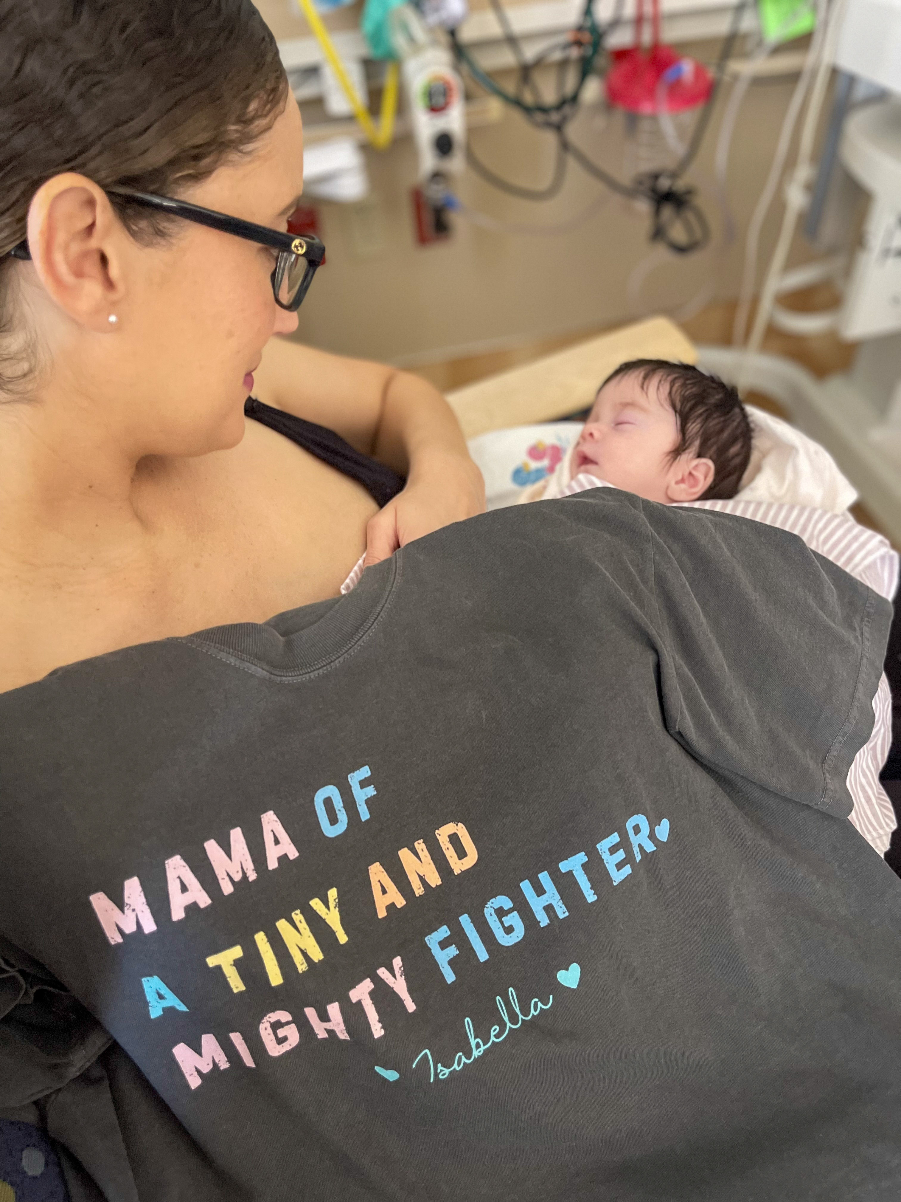 A mother holds her sleeping newborn baby in a hospital setting. She wears a dark T-shirt that reads "Mama of a Tiny and Mighty Fighter – Isabella" in colorful letters.