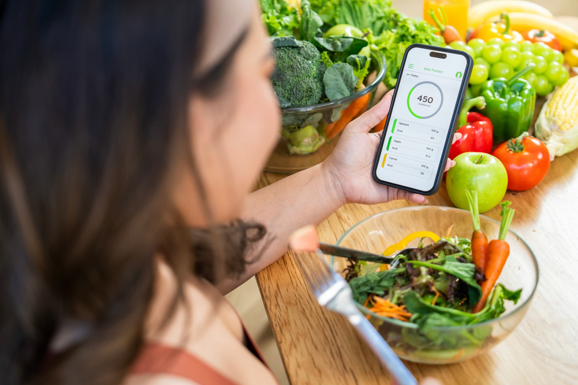Woman checking her dietary progress using a mobile app, while eating a fresh salad with various vegetables.