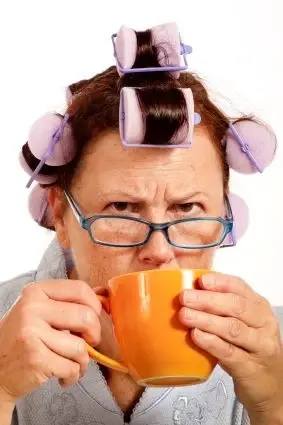 An older woman looking irritated, with hair rollers on her head, while drinking from an orange cup of coffee.