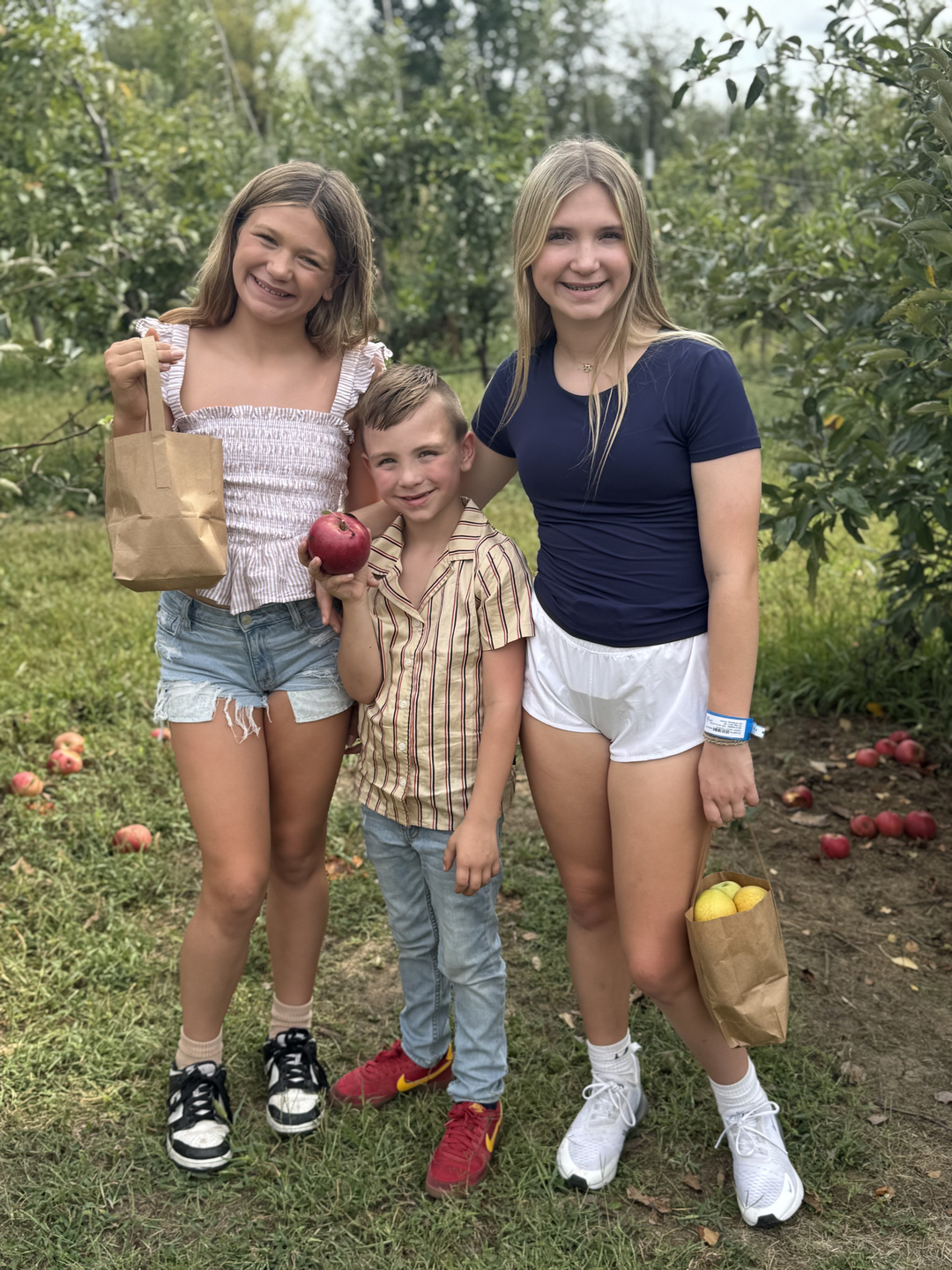 Three children smiling and holding bags of apples in a garden.