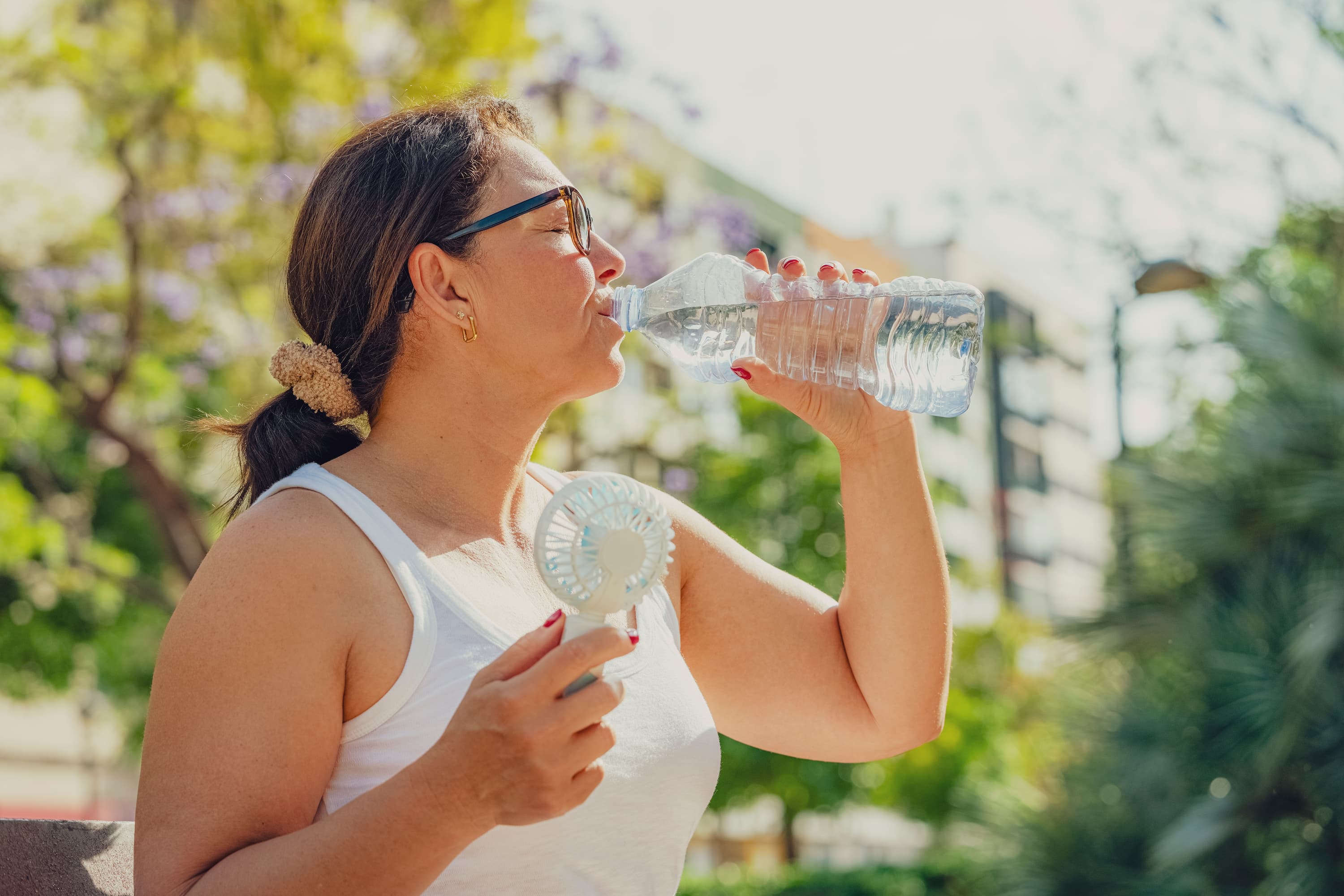 Woman drinking water and holding a handheld fan outdoors on a hot summer day to prevent heat-related illness.