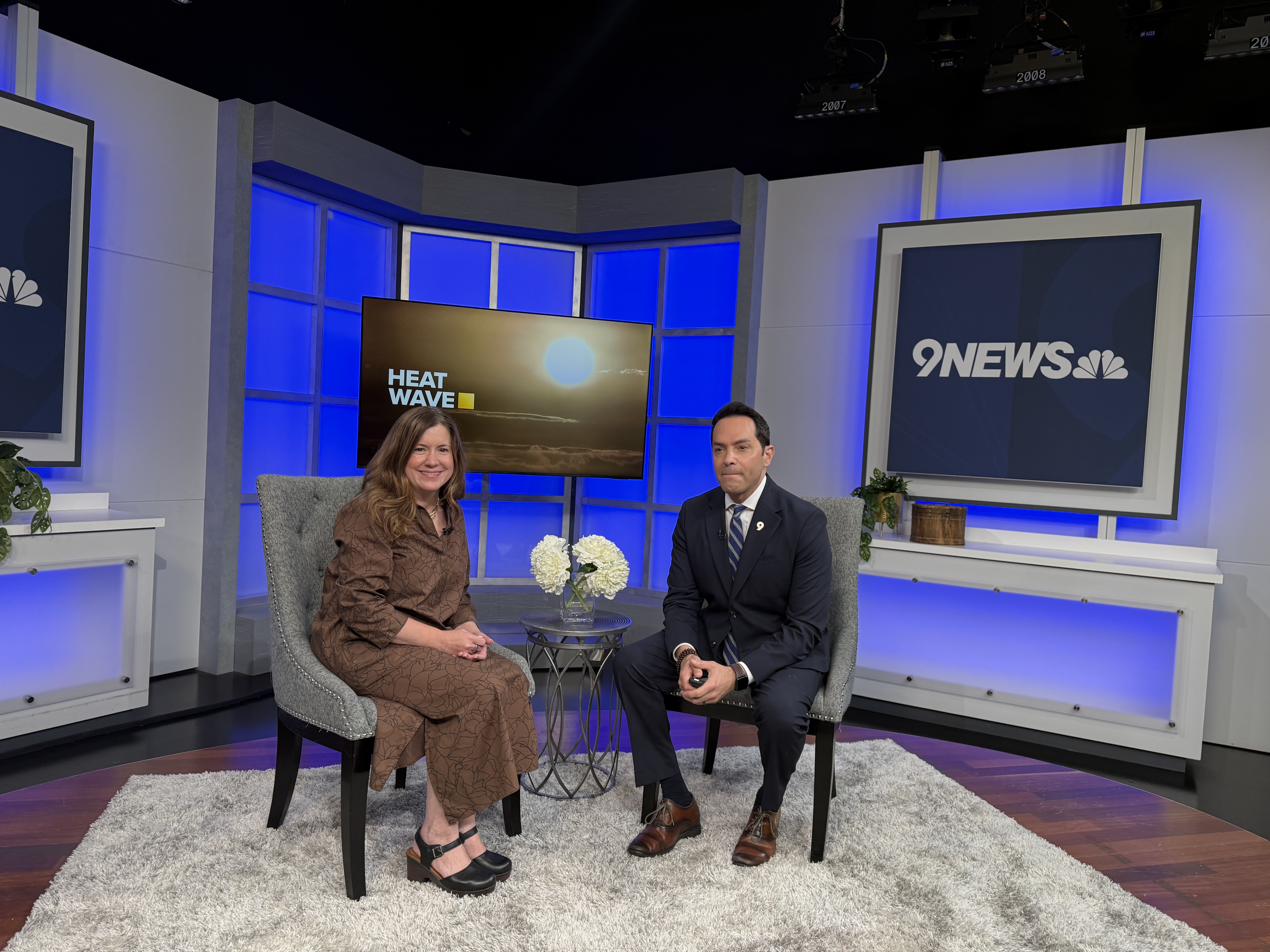 Woman and man seated in chairs in a television studio with screens that say "Heat Wave" and 9 News