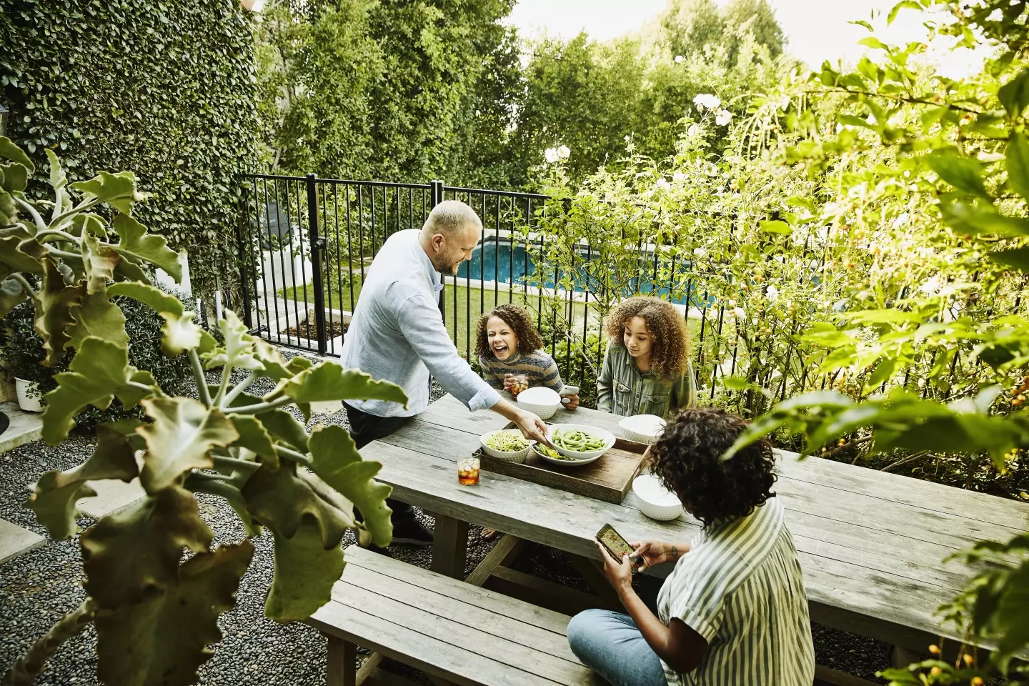 A smiling and laughing family of four shares a meal at a picnic table in a courtyard outside, with a swimming pool in the background.