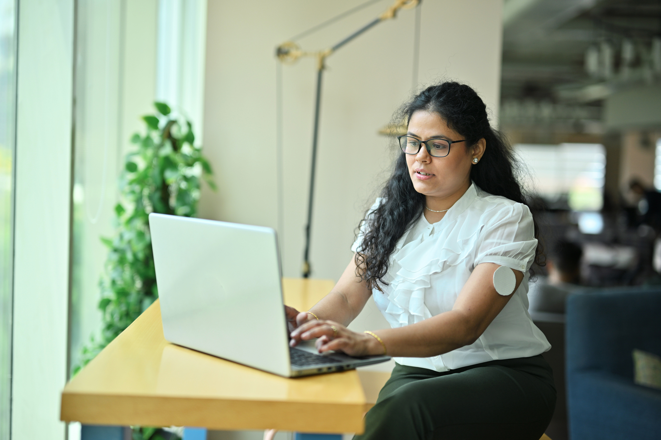 A woman working on laptop in an office space, with a diabetic sensor on her arm.