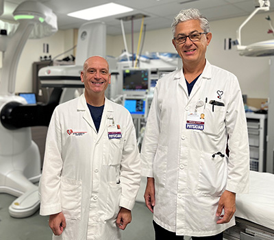  Dr. Niuton Koide and Dr. Erik Sirulnick, pose standing in an operating room, smiling.