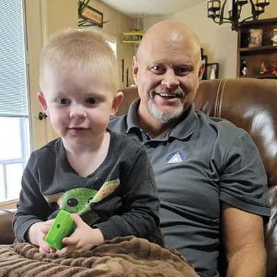 Stephen Gacioch smiles while holding his grandson in their living room.