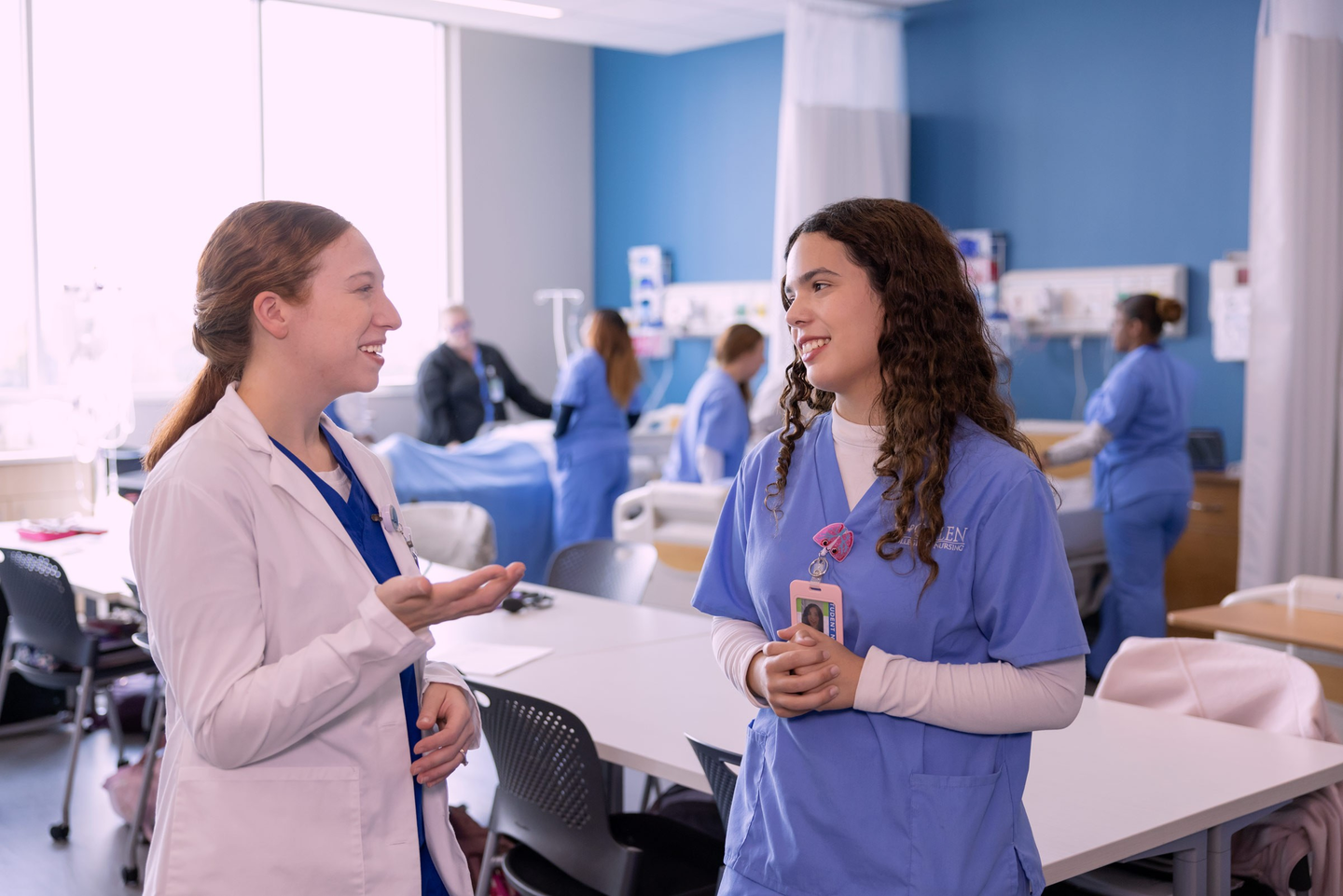 A nursing instructor in a white lab coat speaks with a student in blue scrubs in a bright clinical simulation lab. Both are smiling and engaged in conversation beside classroom tables, while other students practice patient-care skills around hospital beds in the background. Large windows, medical equipment, and blue accent walls create a modern healthcare training environment.