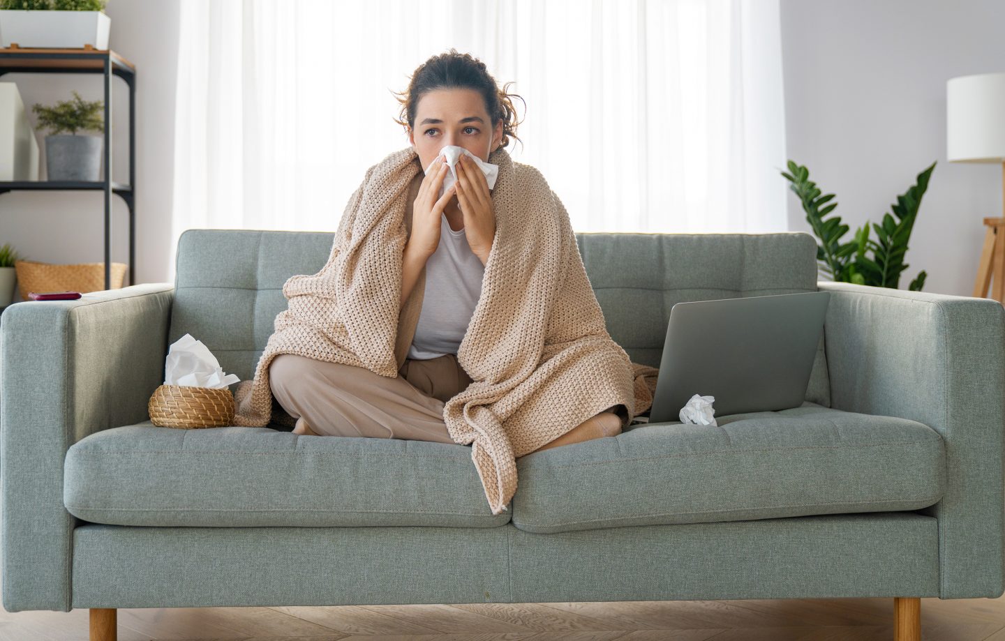 Young woman feeling ill, sits on a couch, while wiping her nose with a tissue.