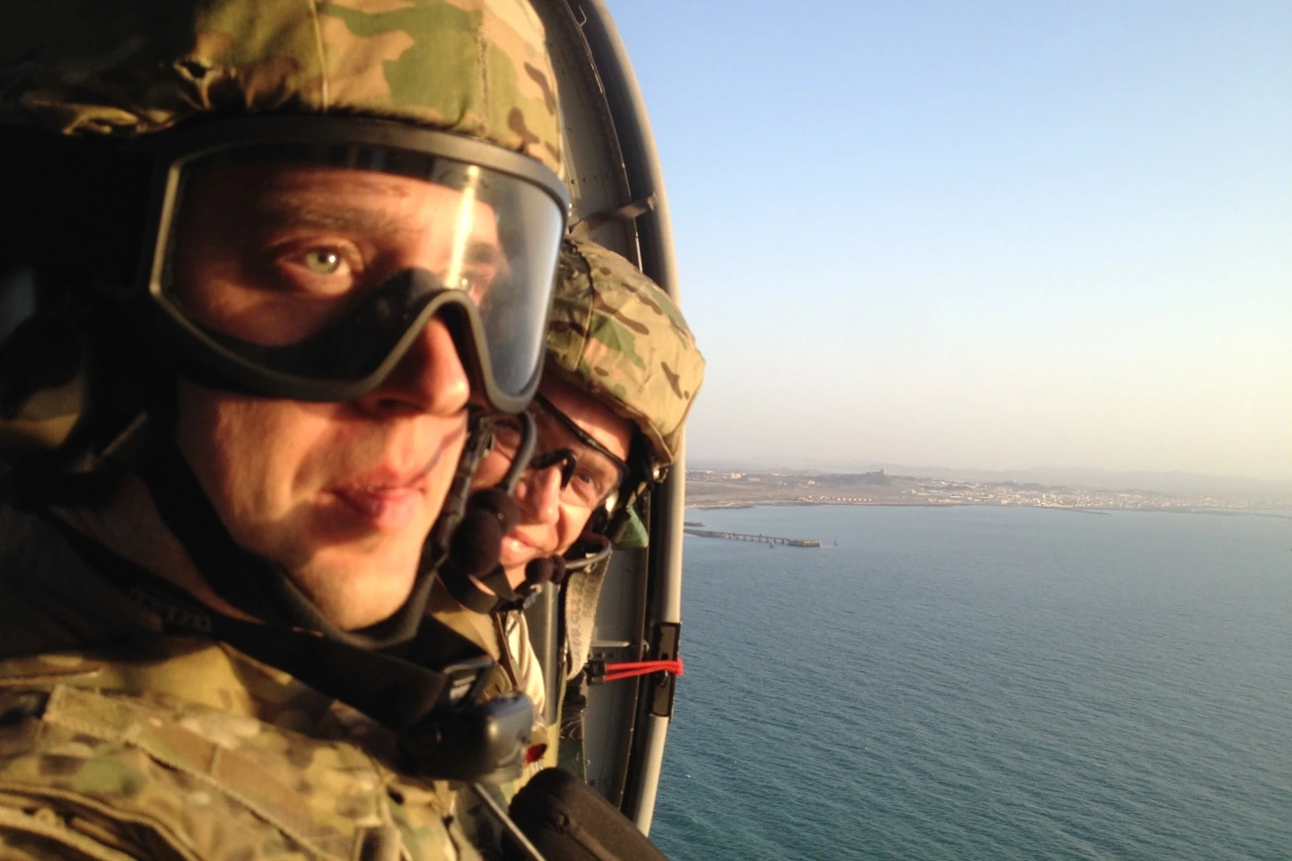 Dr. Michael Rawlins and another Air Force memeber, both wearing camouflage helmets, uniforms, and goggles, looking out of a Blackhawk helicopter over the ocean.