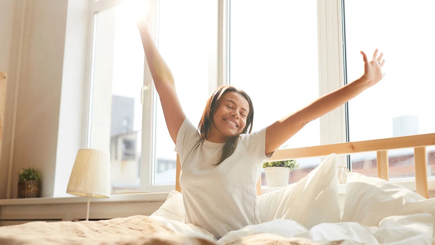 A young woman sitting up in bed, arms outstretched, smiling warmly after waking up.