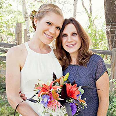 Emily Mosman and her mom hug while standing together in a garden. Emily is holding a bouquet of flowers and is wearing a while dress, while her mom wears a blue blouse.