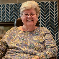 Ada smiles while sitting down, wearing a purple, blue and yellow floral print shirt.