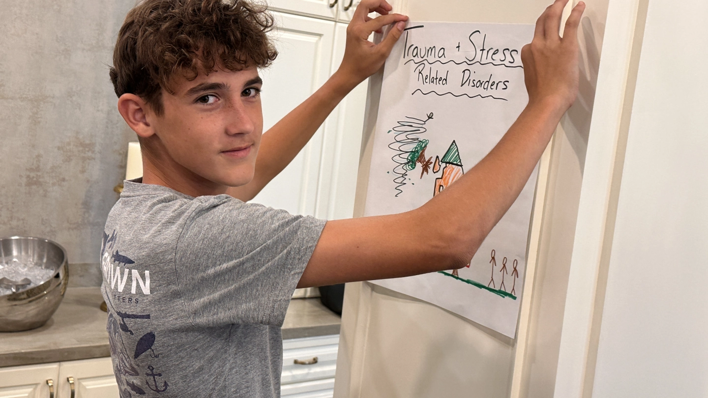 A person hangs a poster titled "Trauma + Stress Related Disorders" on a wall. The poster features a drawing of a house, a tornado, and stick figures. The setting appears to be a kitchen, with cabinets and countertops visible in the background.
