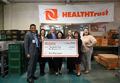 The Good Samaritan Hospital Canned Food Drive Committee presenting an oversized $10,000 check to The Health Trust CEO Michele Lew, in a food drive collection center, with a large white banner reading 'HealthTrust' above them.
