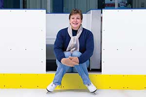 Denise Dwinnell wearing ice skates sitting on the side of an ice rink