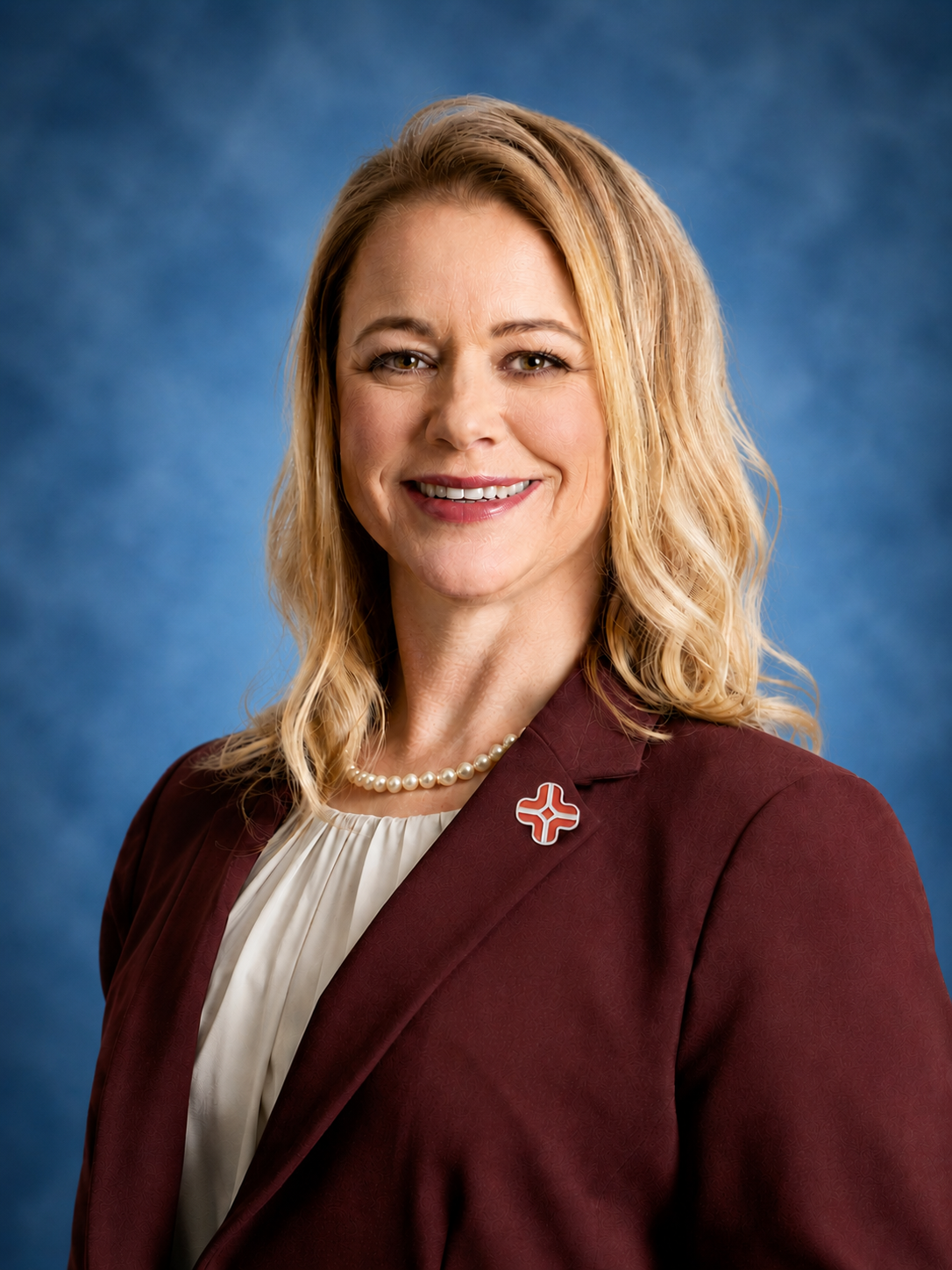 Professional headshot of a person wearing a burgundy blazer, white blouse, and pearl necklace against a blue studio background.