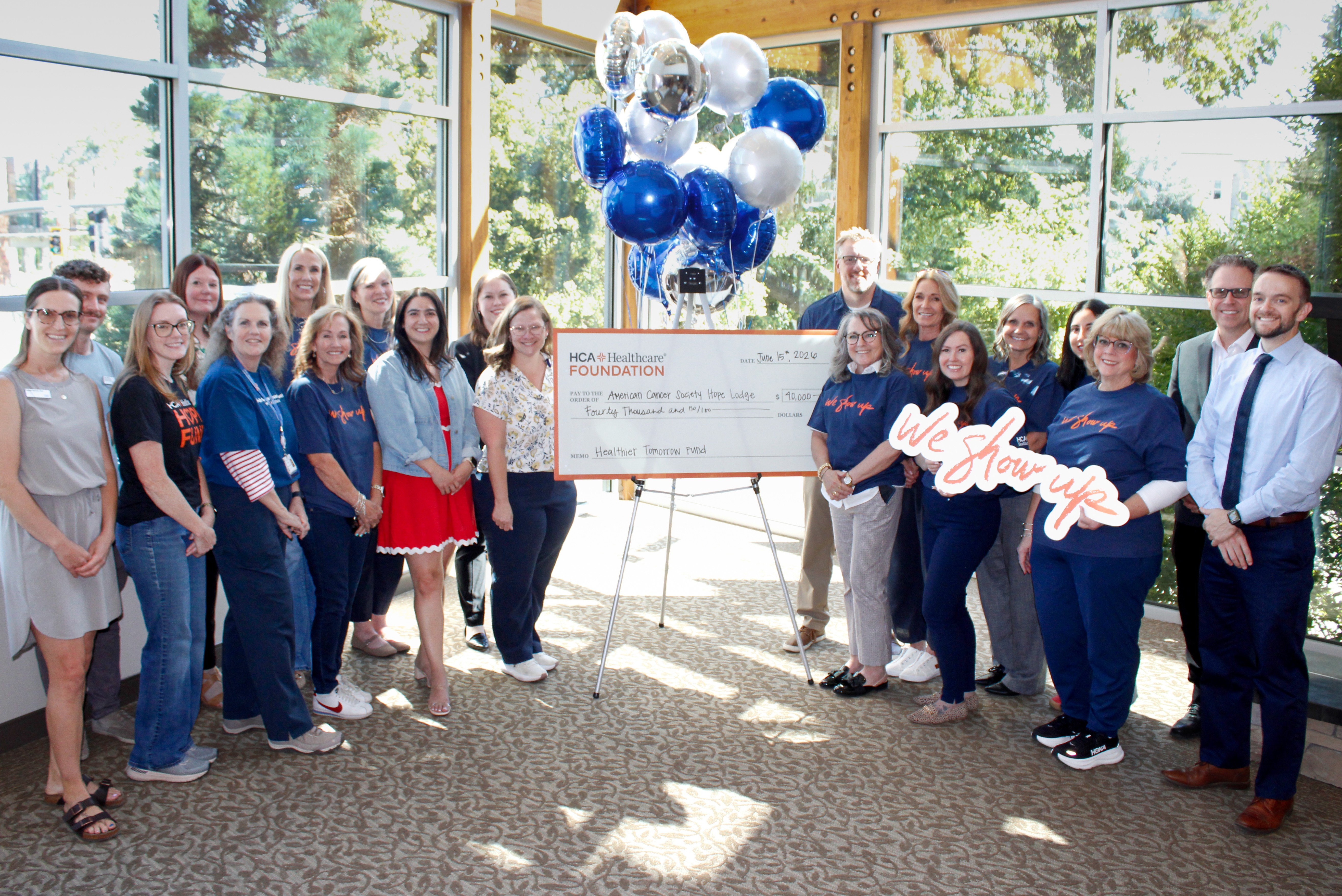 A group of approximately 20 representatives from MountainStar Healthcare, the HCA Healthcare Foundation, and the American Cancer Society Hope Lodge gather indoors around a ceremonial $40,000 grant presentation check. The group stands beneath blue, white, and silver balloons in a bright room with large windows and views of trees outside. Several participants wear blue "We Show Up" shirts, while others are dressed in business attire. The oversized check recognizes funding from the HCA Healthcare Foundation to support the American Cancer Society Hope Lodge in Salt Lake City.