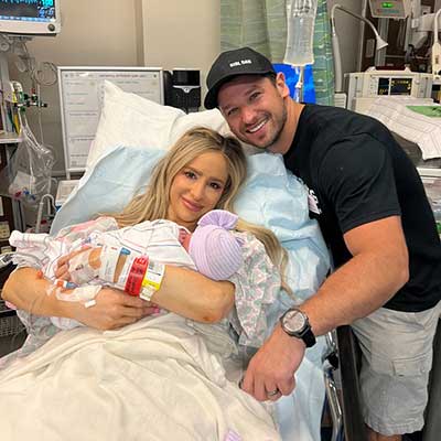 Katie Corwin in a hospital bed smiles with her newborn baby in her arms and her husband at her side.